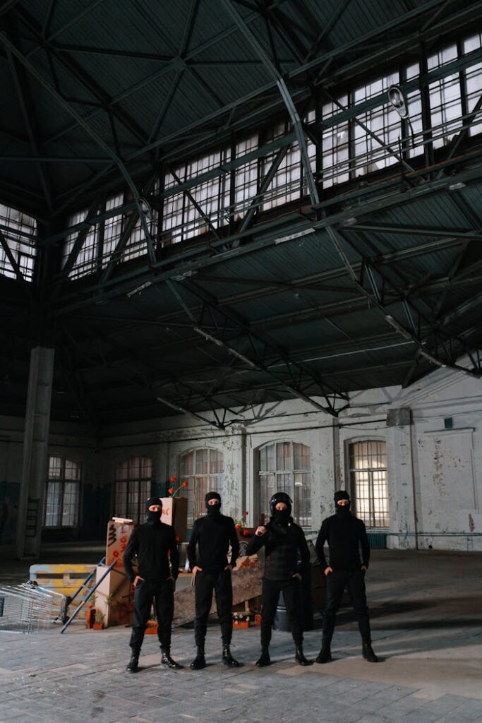 A group of masked activists stands united in a spacious, abandoned warehouse setting.